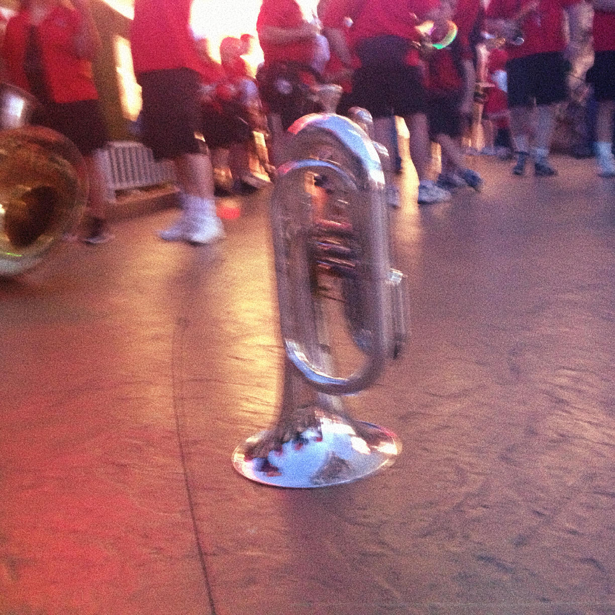 A marching baritone horn sitting on the ground. In the background are musicians in red shirts and black shorts.